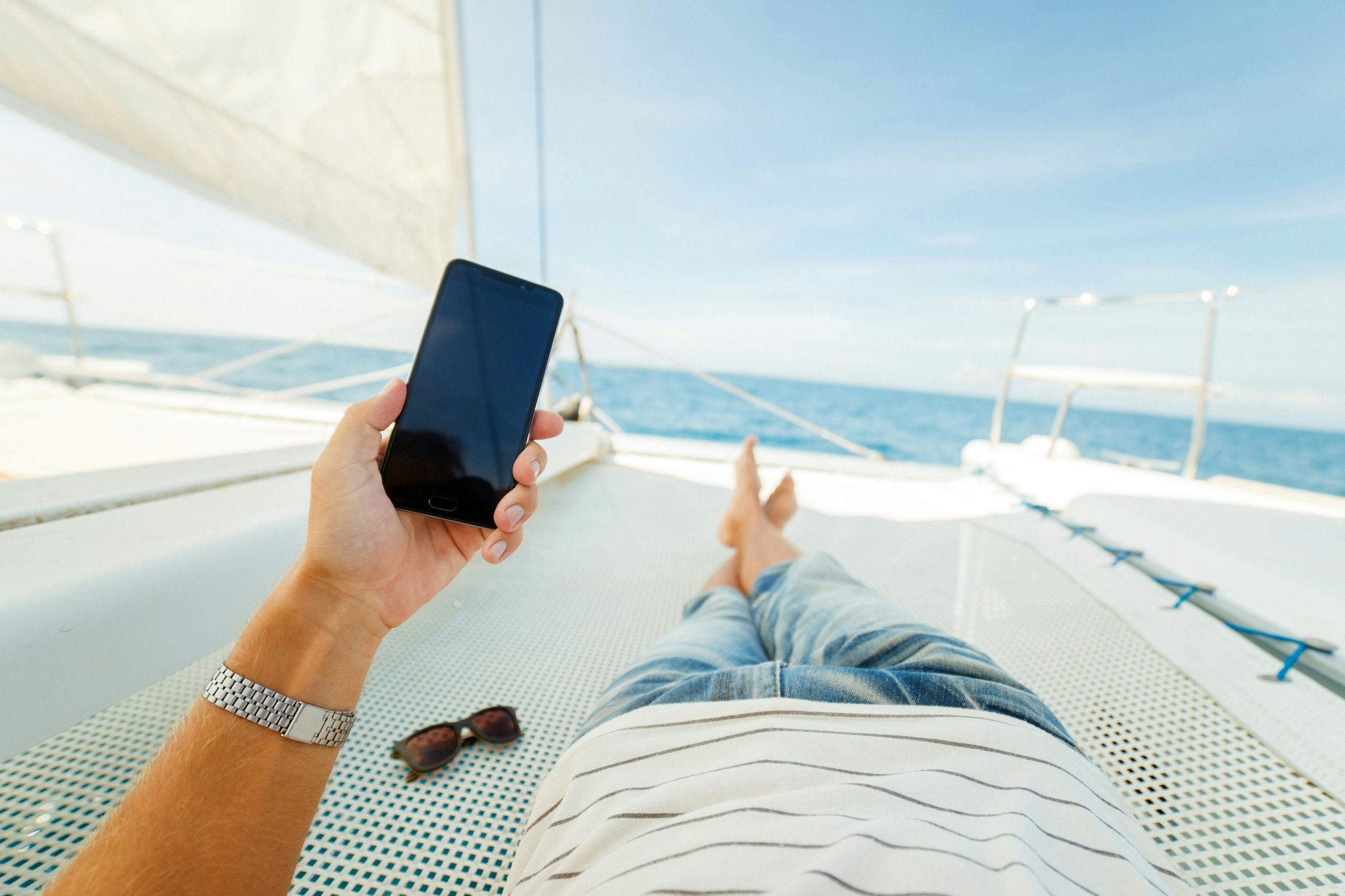 Mann slapper av på en seilbåt med mobilen i hånden, bildet er tatt fra personen sitt perspektiv