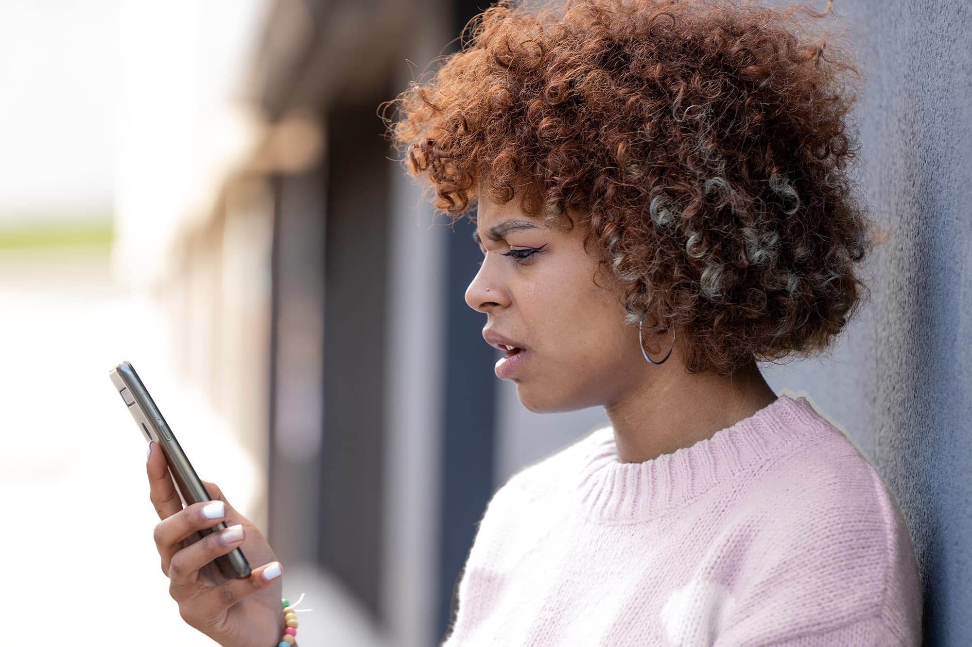 En bekymret kvinne med krøllete hår og en lys rosa genser ser forvirret og frustrert ut mens hun stirrer på mobiltelefonen sin. Hun står utendørs ved siden av en grå vegg på en solfylt dag. Hennes uttrykk tyder på at hun kan ha mottatt en urovekkende eller forvirrende melding eller anrop.