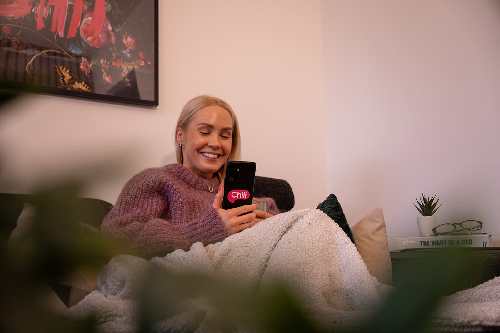 En smilende kvinne sitter komfortabelt i en sofa under et lyst pledd, iført en tykk, lilla genser. Hun holder en mobiltelefon med Chilimobil-logoen tydelig synlig på skjermen. I bakgrunnen sees en plante, en bokhylle med bøker og briller, samt en dekorativ plakat på veggen. Stemningen er avslappet og hjemmekoselig.