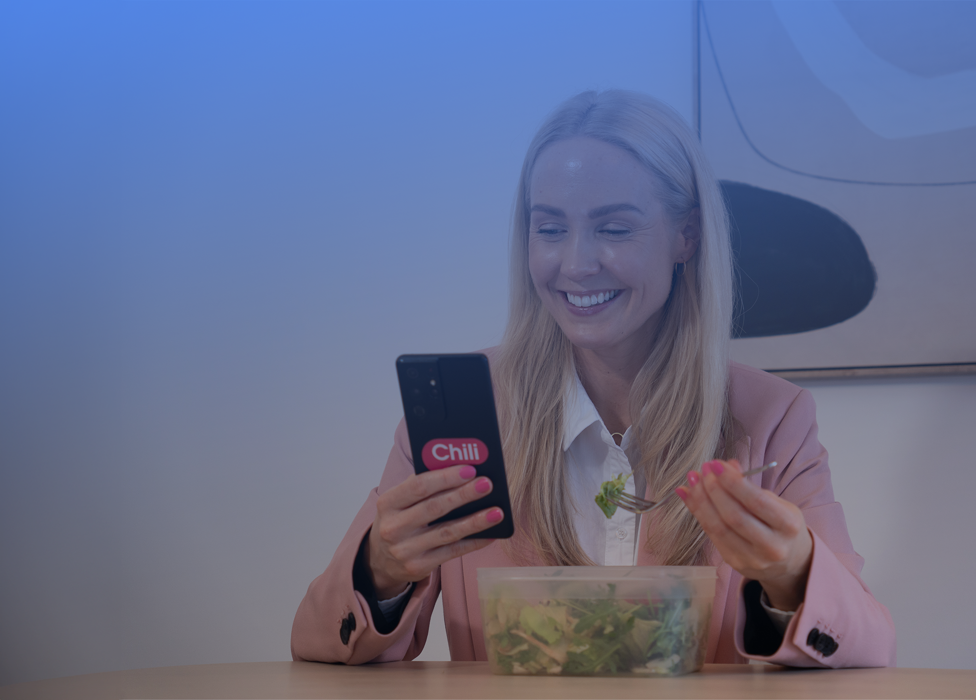 En smilende kvinne som holder en mobiltelefon med en logo som sier "Chili". Hun har blondt hår, har på seg en lys rosa blazer over en hvit skjorte, og spiser salat fra en klar plastboks. Kvinnen er inne, med en moderne kunstig bakgrunn som inneholder myke blå toner og en abstrakt, mørk form. Atmosfæren er avslappet og moderne.