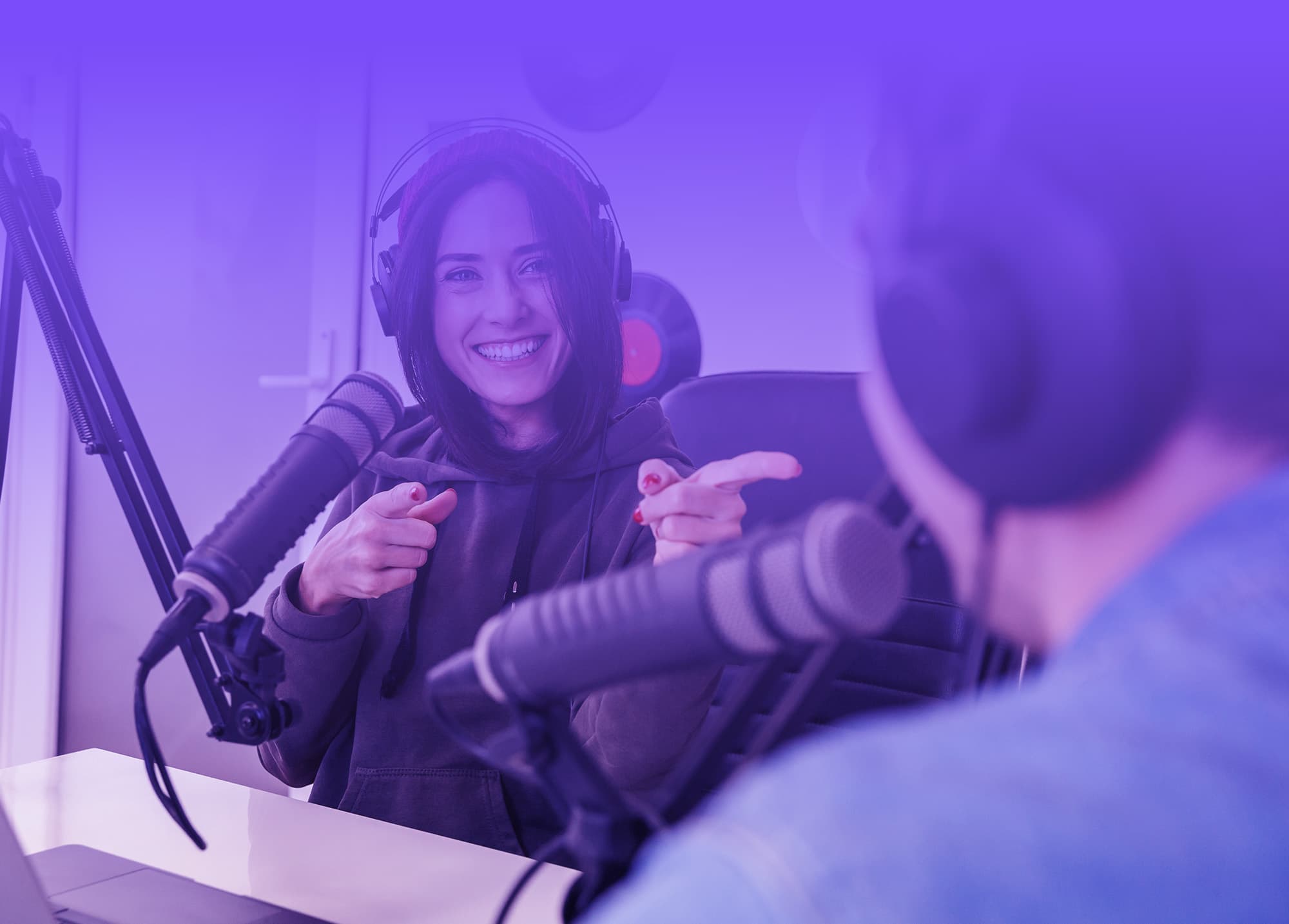 en kvinne og en mann som sitter i et podcast-studio. Kvinnen, som er i forgrunnen og ser inn i kameraet, smiler bredt og peker mot mannen med begge hender. Hun har på seg hodetelefoner og en mørk hettegenser. Mannen, som er delvis ute av fokus og med ryggen til kameraet, sitter foran en mikrofon, lik kvinnen. Studioet har et monokromatisk lilla lys som gir en moderne og kreativ atmosfære. De ser ut til å ha en avslappet og vennlig samtale. Belysningen og fargetonene i bildet er konsistent, og gir en følelse av samhørighet og fokus på dialogen som foregår.