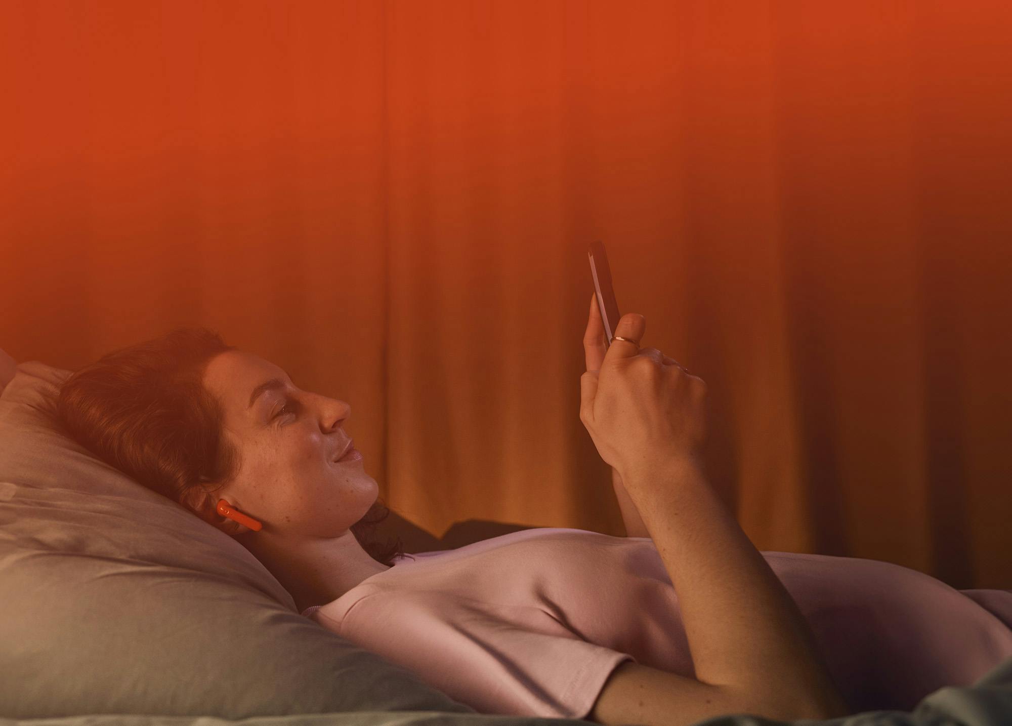 En kvinne ligger behagelig på ryggen i en seng med hodet på en pute. Hun smiler og ser på en smarttelefon hun holder i hånden. Kvinnen har oransje ørepropper, og rommet er opplyst i en varm oransje fargetone, noe som skaper en avslappet og koselig atmosfære. Den rolige scenen antyder at hun lytter til en lydbok eller nyter multimedia-innhold på sin enhet.
