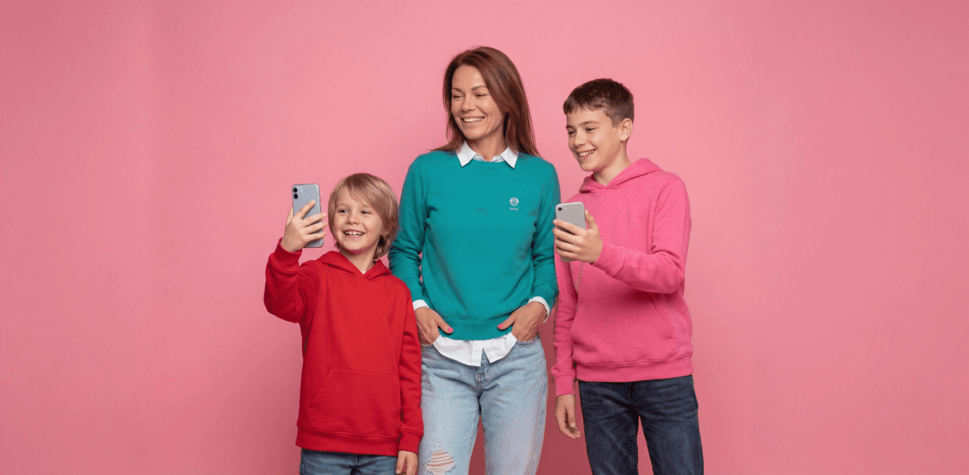 Smilende kvinne i grønn genser står mellom to gutter som begge holder mobiltelefoner. Den yngste gutten til venstre har rød hettegenser, mens den eldre gutten til høyre har rosa hettegenser. Bakgrunnen er ensfarget rosa, og stemningen er lys og uformell.