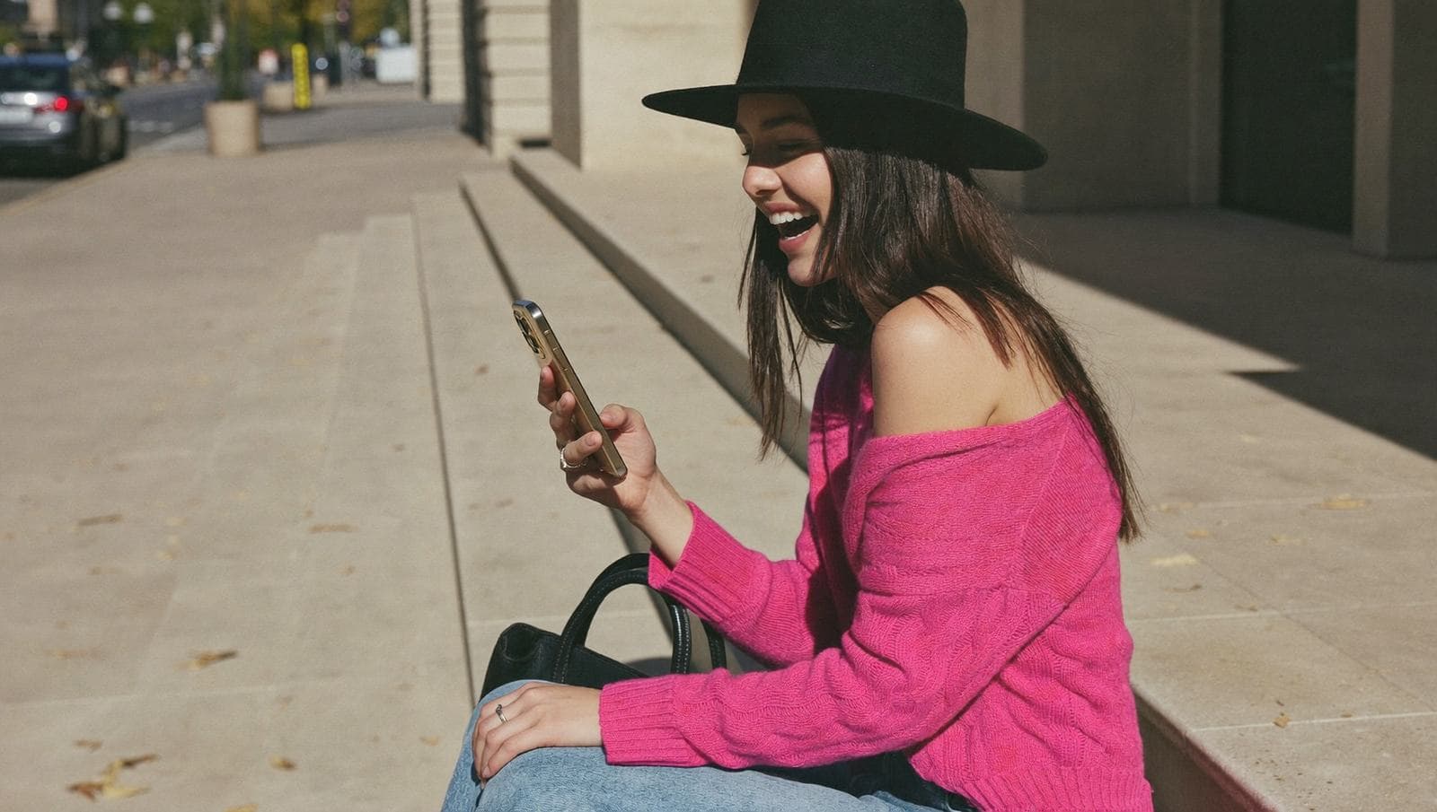 Kvinne i rosa genser og svart hatt sitter på ei trapp i byen og ler av noe på mobilen.